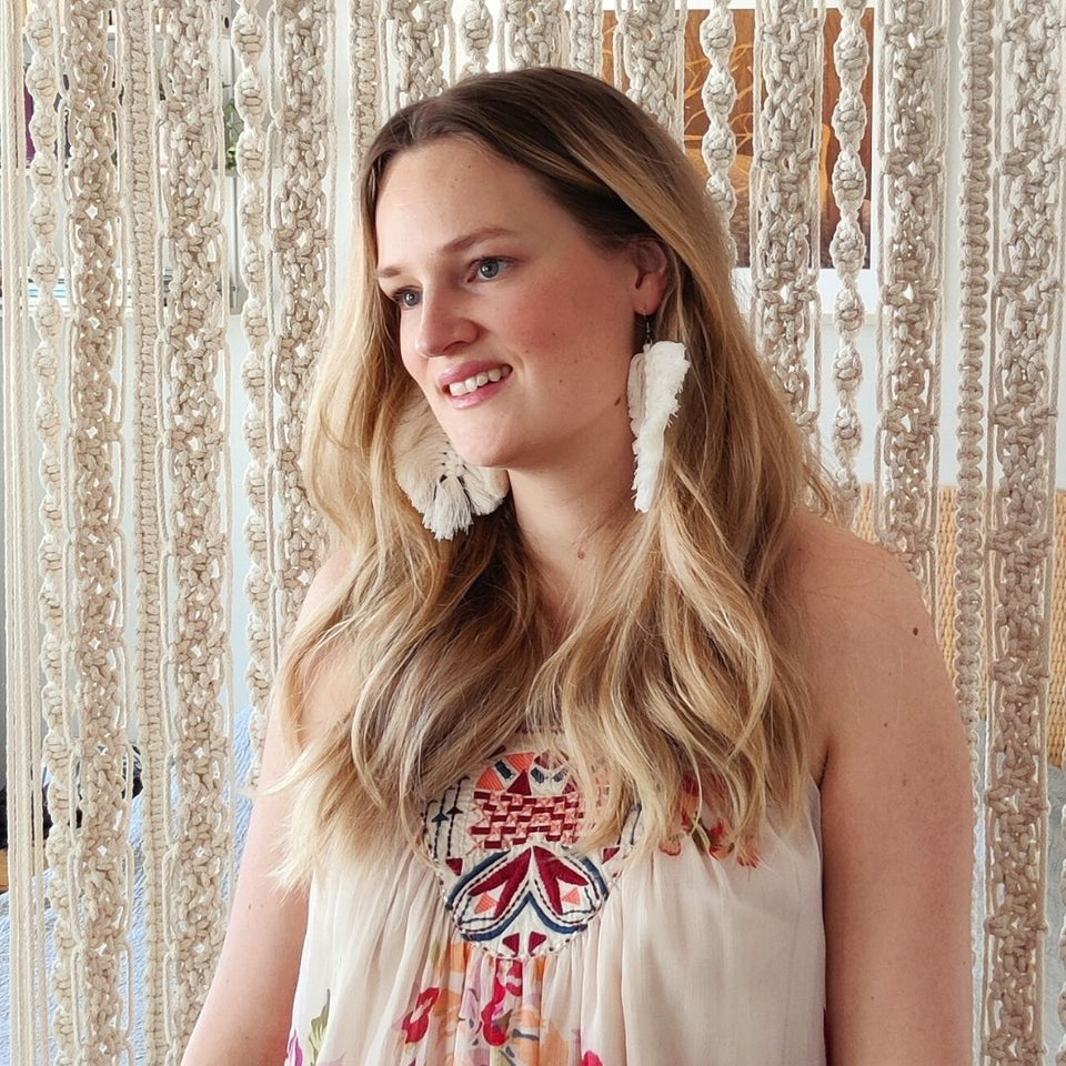 Macramé maker standing in front of the macramé curtain, wearing white macramé feather earrings and a patterned light shirt.