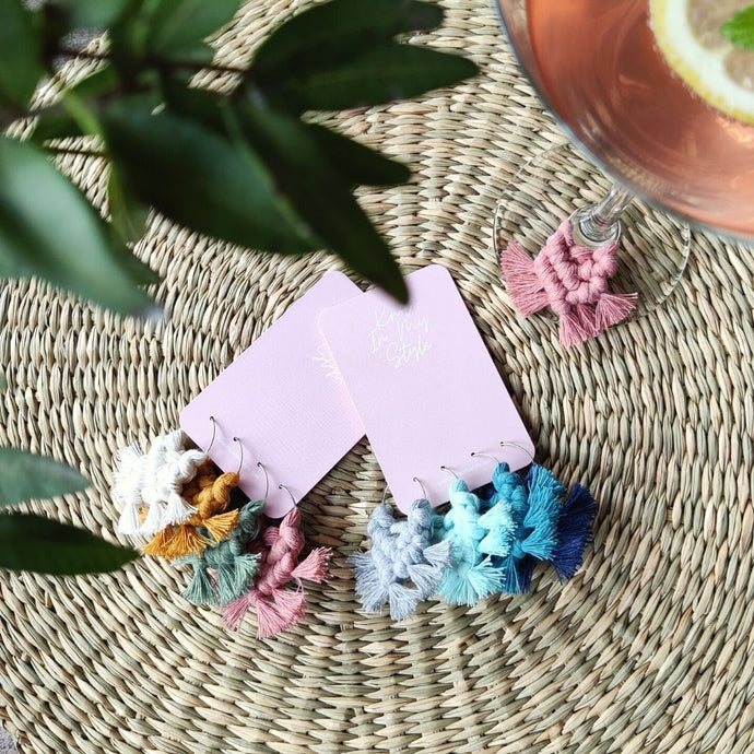 Two sets of macramé My Precious Wineglass Charms in shades Garden Party and Ocean View. On the top right corner is a pink drink with a lemon slice, green leaves on the left side of the picture.