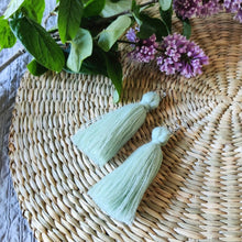 Load image into Gallery viewer, Macramé tassel earrings Pillow Talk in shade Mint Green laying flat on a straw place mat, lilacs on the side.
